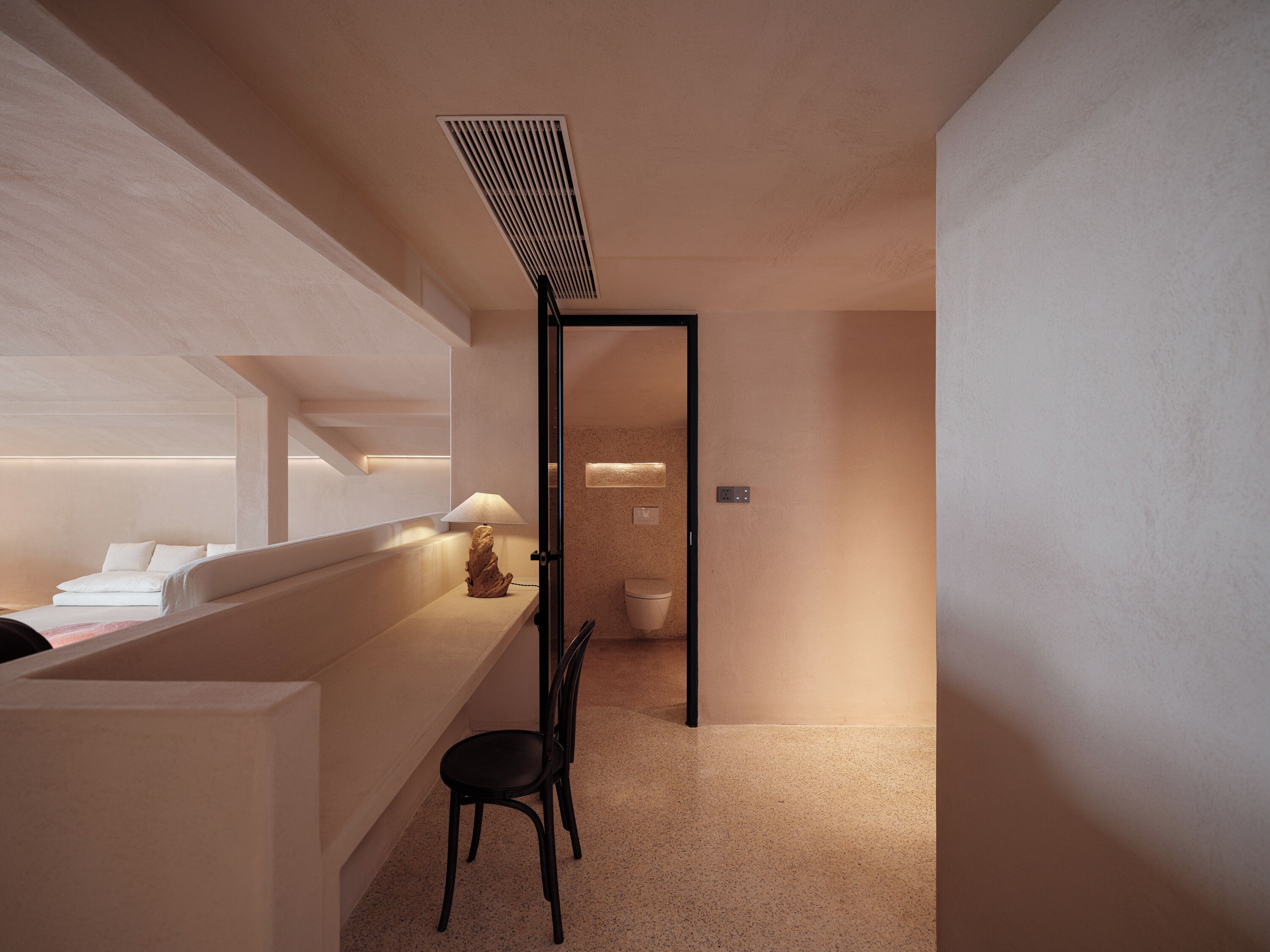 A modern hotel room with a minimalist design, featuring a desk with a lamp, a chair, and a bed.