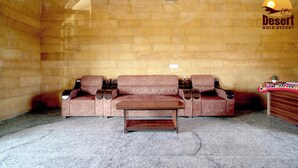 A room with a beige brick wall, a brown sofa, a wooden coffee table, and a small wooden side table with a decorative item. - Desert Gold Resort (Jaisalmer)
