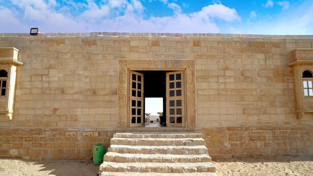A stone building with a large open doorway, steps leading up to it, and a green barrel on the ground. - Desert Gold Resort (Jaisalmer)