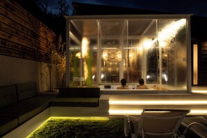 Un'area spa moderna all'aperto con struttura in vetro, una zona relax con panchina imbottita e una piccola piscina.