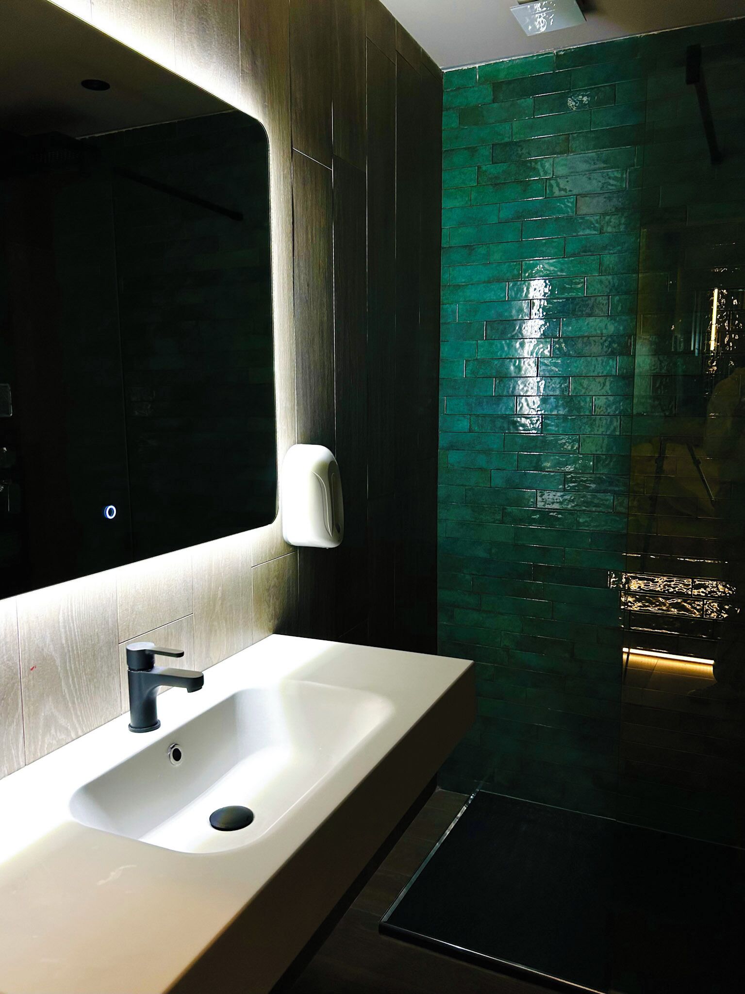 Un baño moderno con un lavamanos blanco, un grifo negro y una cabina de ducha con mampara de vidrio verde.