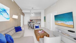 Un salon moderne avec un canapé, une table basse et un téléviseur. On aperçoit à l’arrière-plan un coin repas et une cuisine.