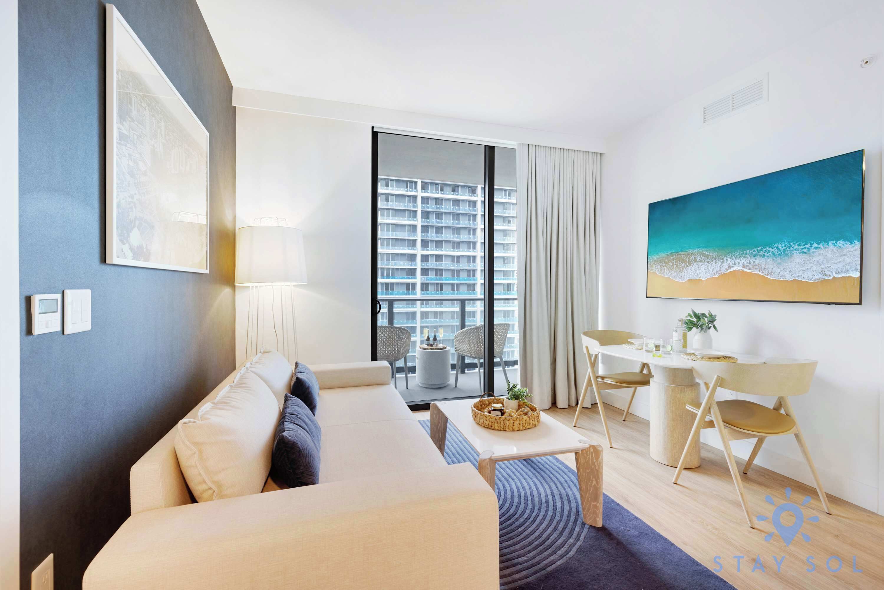 A modern living room with a sofa, a dining table with chairs, a large window with a view of a building, and a painting of a beach.