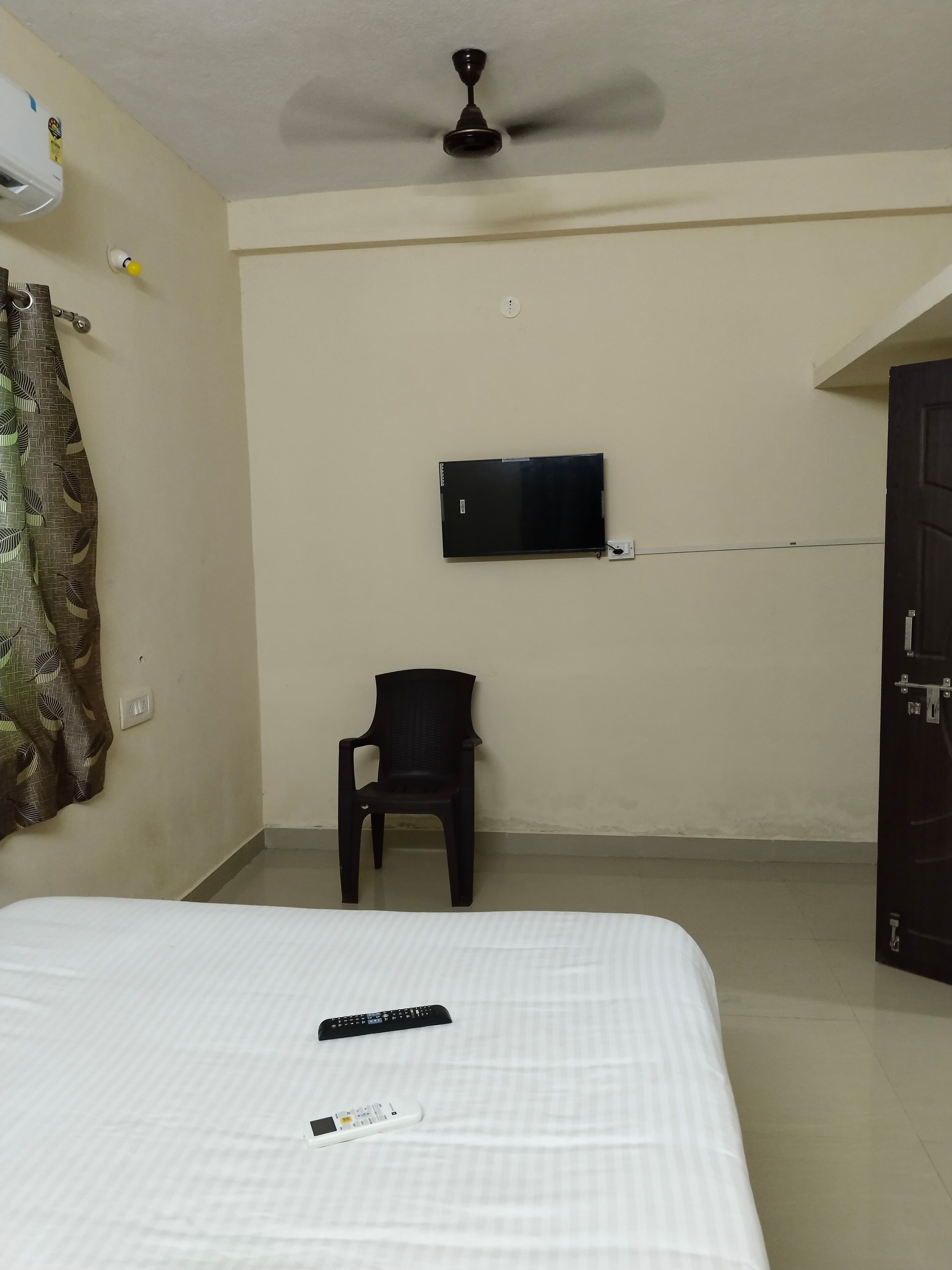 A hotel room with a bed, a chair, a TV mounted on the wall, and a ceiling fan.