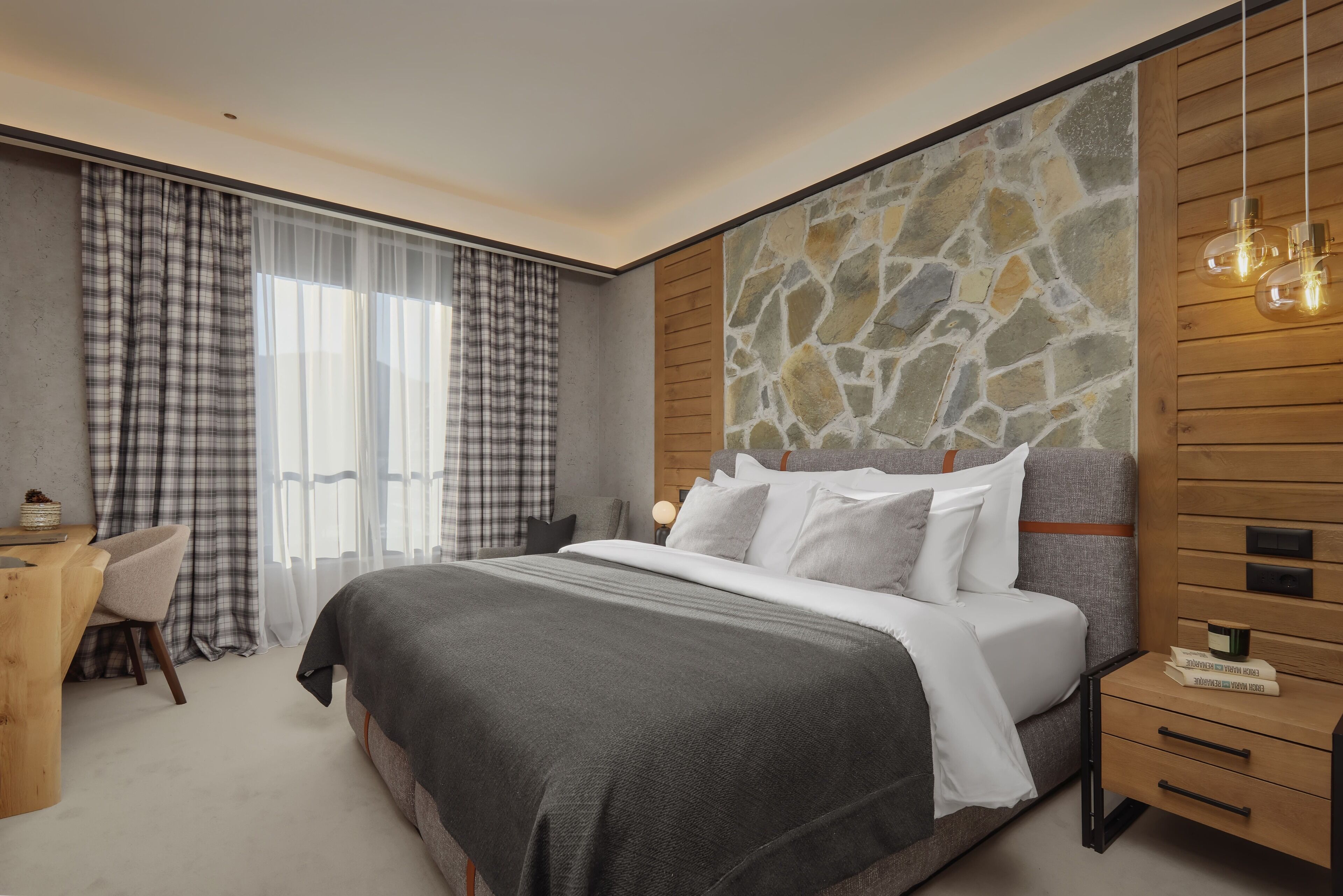 A modern bedroom with a stone accent wall, a large bed with white bedding, a wooden nightstand, and a desk with a chair.