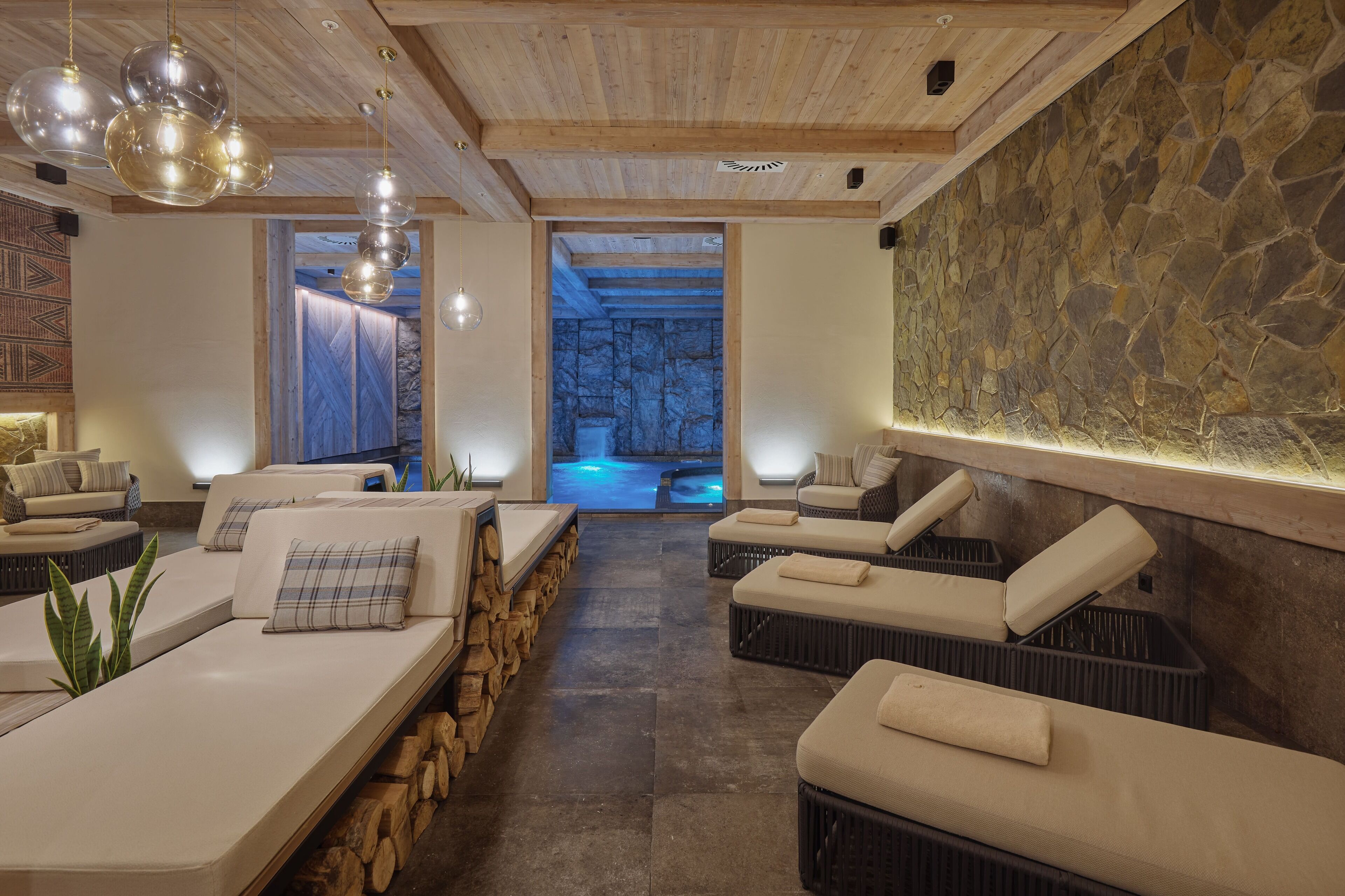 A spa room with two seating areas, wooden beams, stone walls, and a stone wall with a blue-lit background.