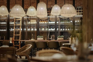 A restaurant interior with multiple tables and chairs, a wine rack in the background, and hanging wicker lamps.