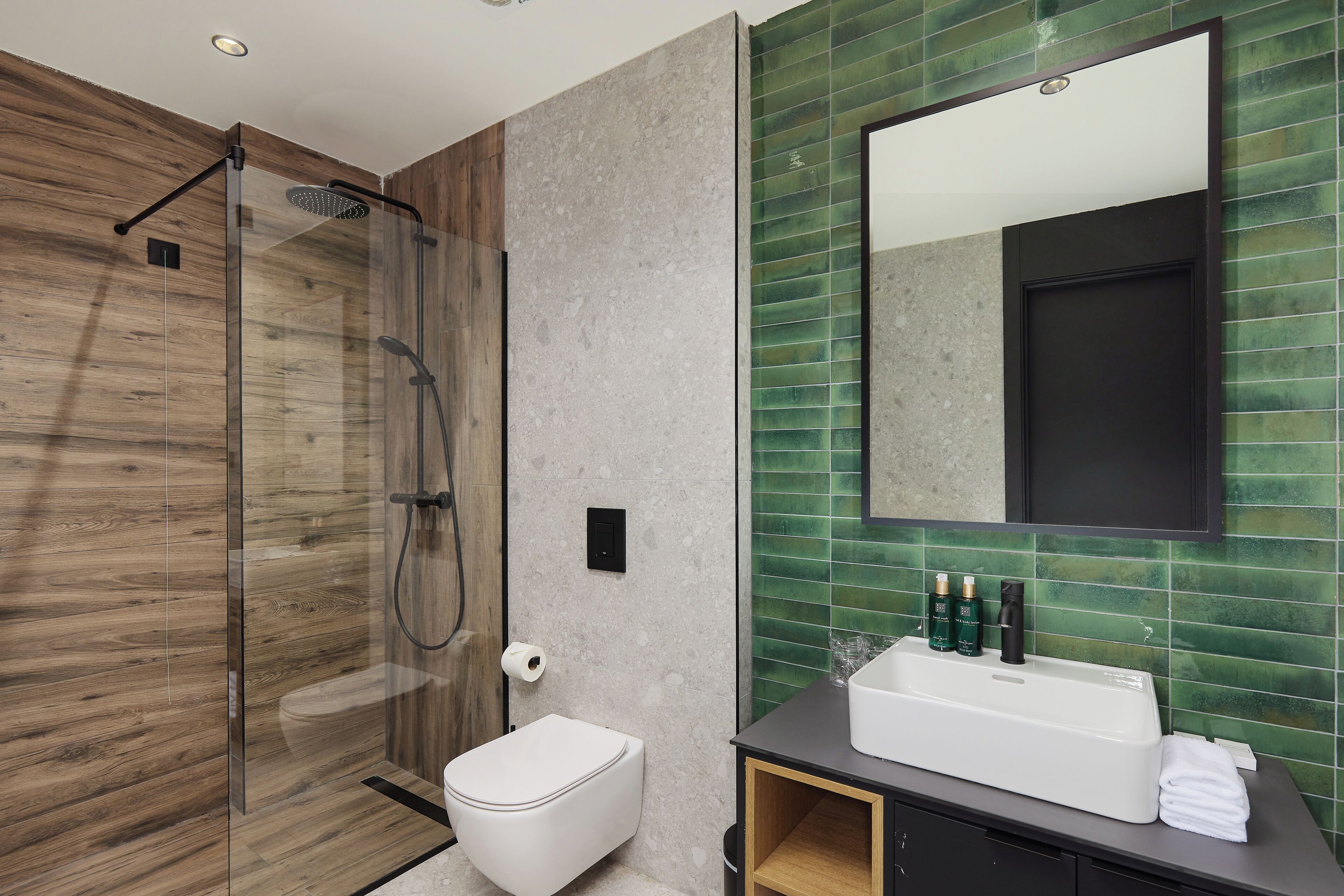 Ein modernes Badezimmer mit einer Duschkabine, einer weißen Toilette, einem großzügigen Waschbecken mit Pflegeprodukten und einer grünen Fliesenwand.