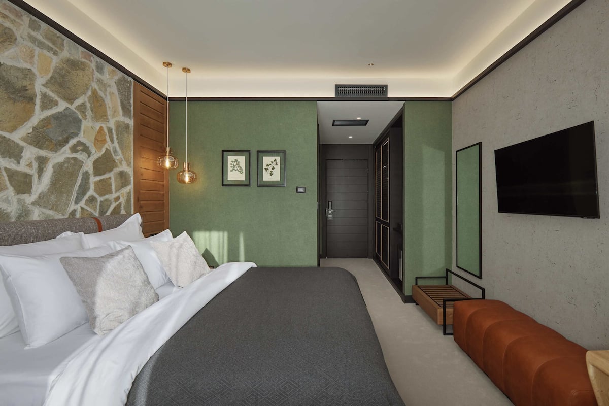 A modern bedroom with a stone accent wall, a large bed with white linens, a wooden bench, and a flat-screen TV.