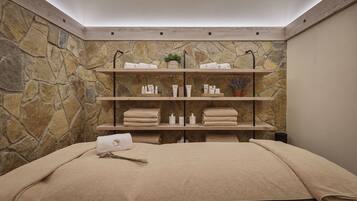 Una stanza spa con parete in pietra, mensole con asciugamani e articoli da toilette, e un letto con coperta beige.