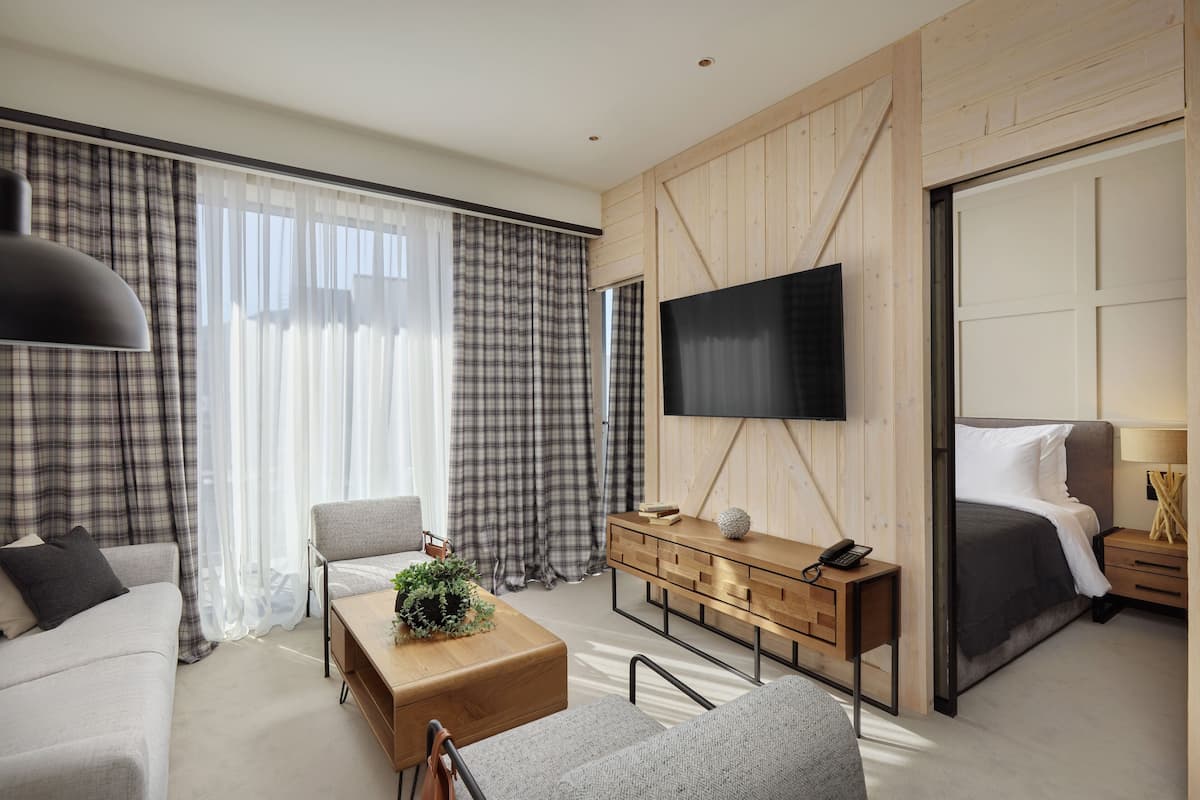 A modern hotel room with a large bed, a TV mounted on the wall, a wooden console table, and a seating area with a sofa and armchair.
