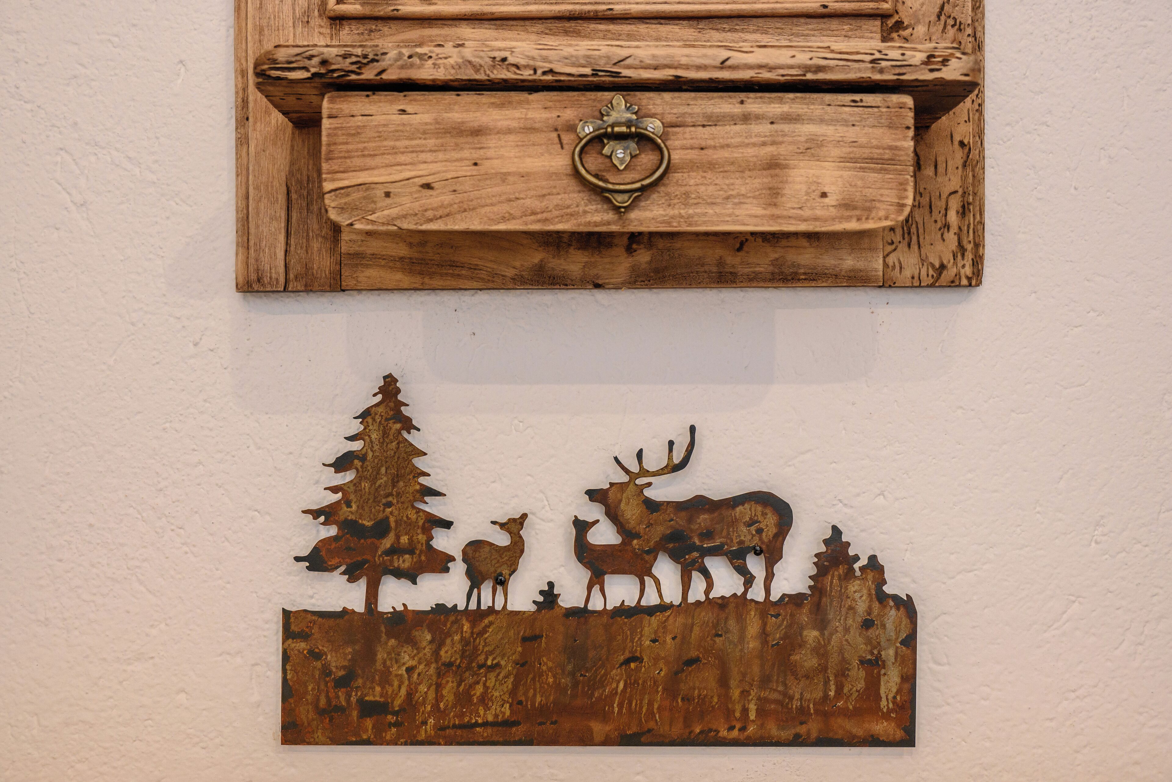 Un meuble rustique en bois, doté d’une poignée en laiton et orné, sur le mur en dessous, d’une silhouette métallique représentant un cerf et un arbre.