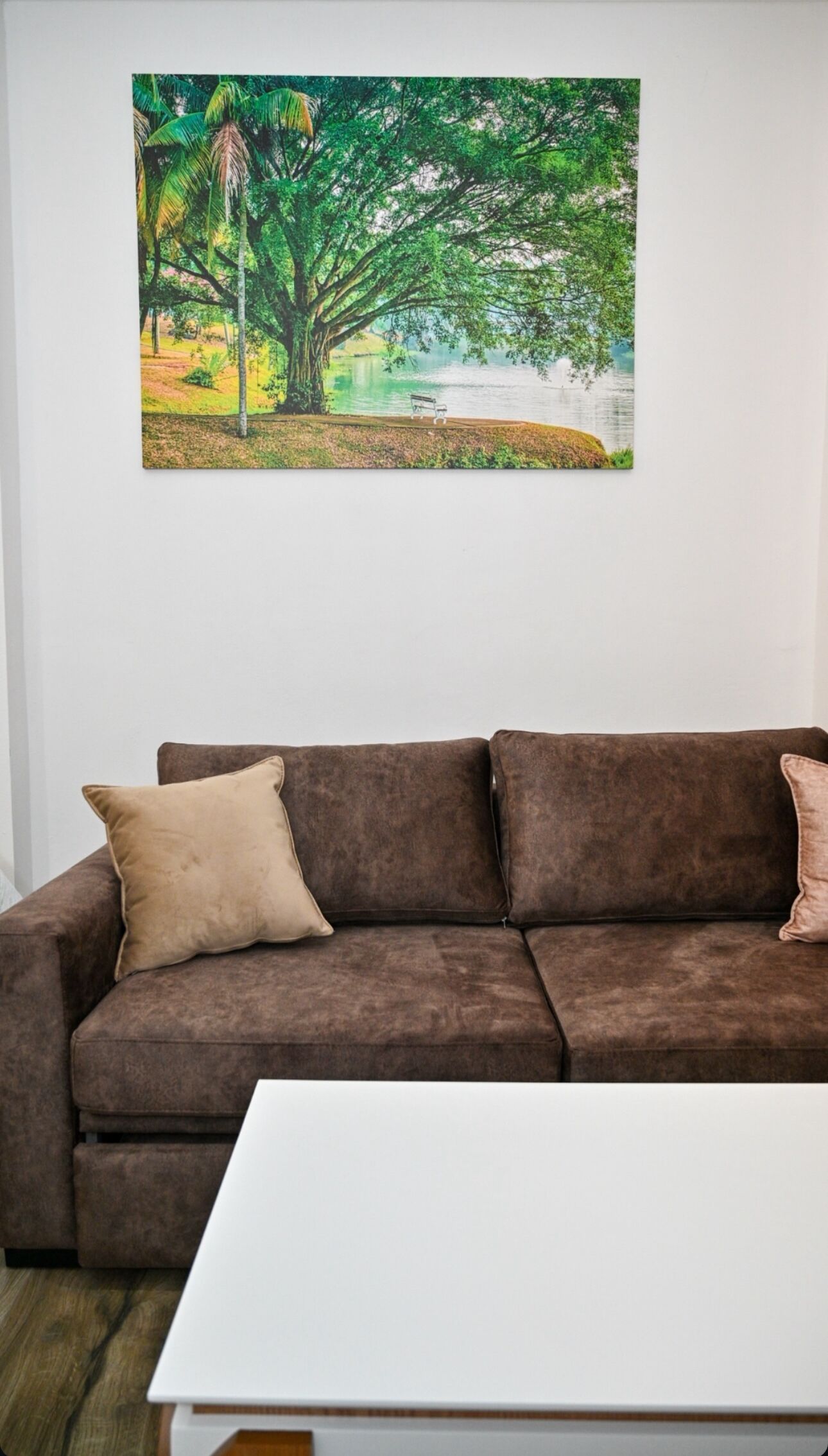 A brown sofa with two cushions, a white coffee table, and a painting of a tree by a lake.