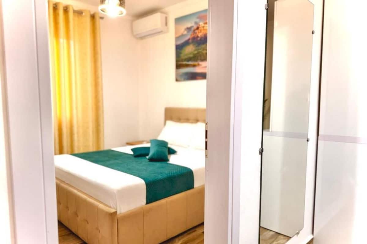 Un dormitorio con una cama, una manta verde, suelo de madera, un cuadro en la pared y una puerta blanca.
