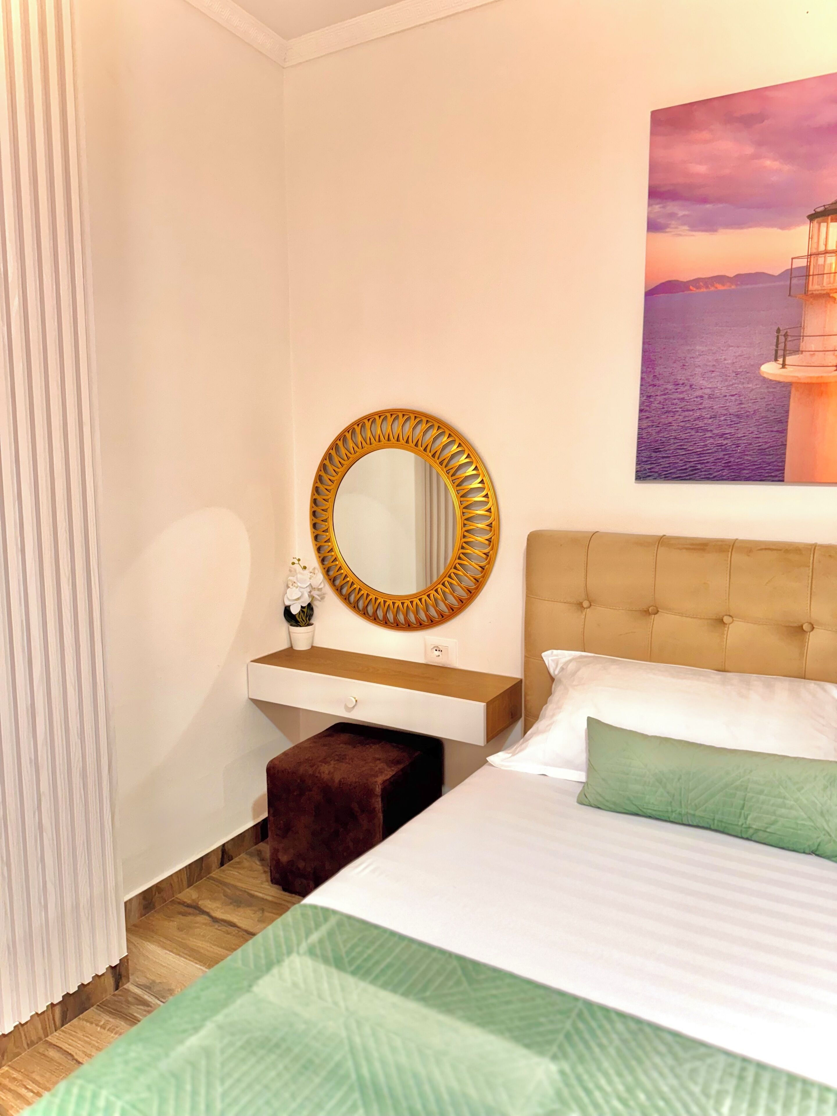 A bedroom with a bed, a bedside table with a mirror, a small stool, and a painting of a lighthouse on the wall.