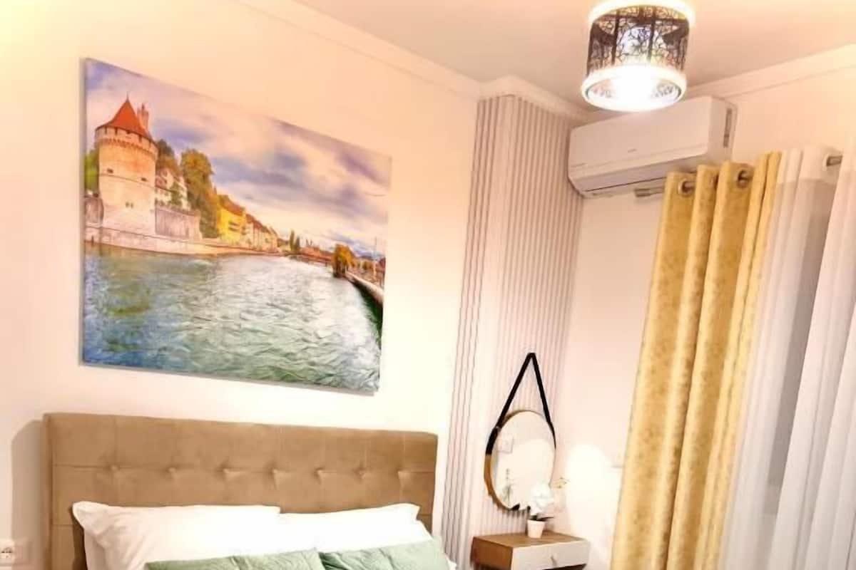 Una cama bien hecha con cabecera, una mesita de noche con lámpara, un cuadro de un castillo y una ventana con cortinas.