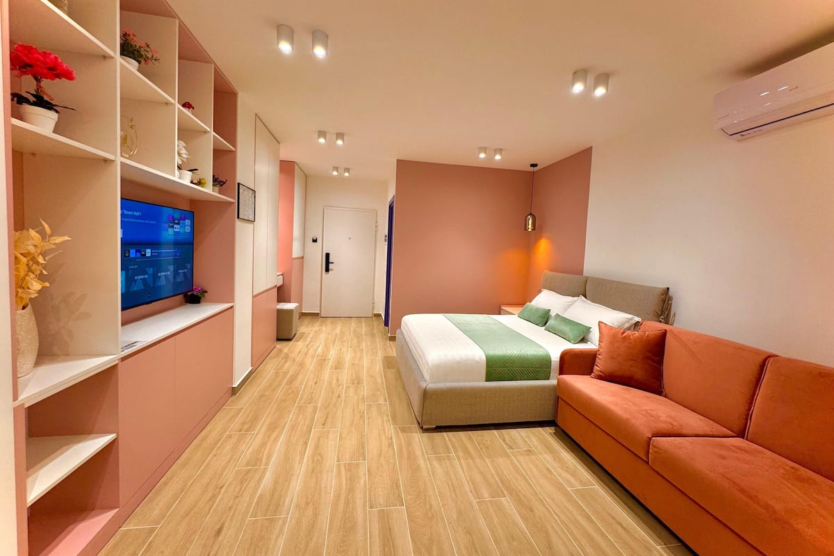 Una habitación de hotel moderna con cama, sofá, televisor en una estantería y una puerta.