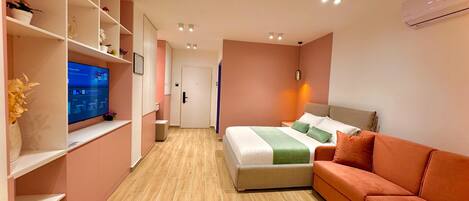 A modern hotel room with a bed, a sofa, a TV on a shelf, and a door.