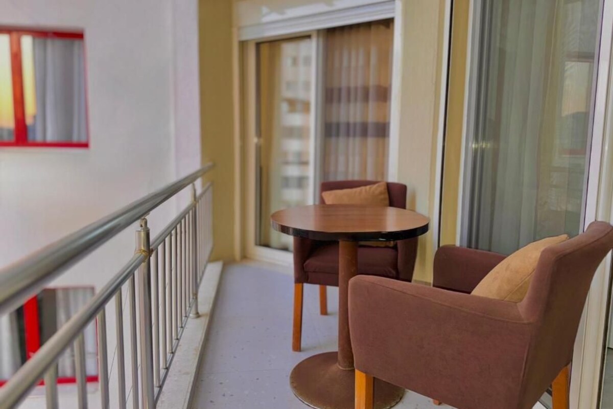 A balcony with a round table and two chairs, a railing, and a view of a building through a glass door.