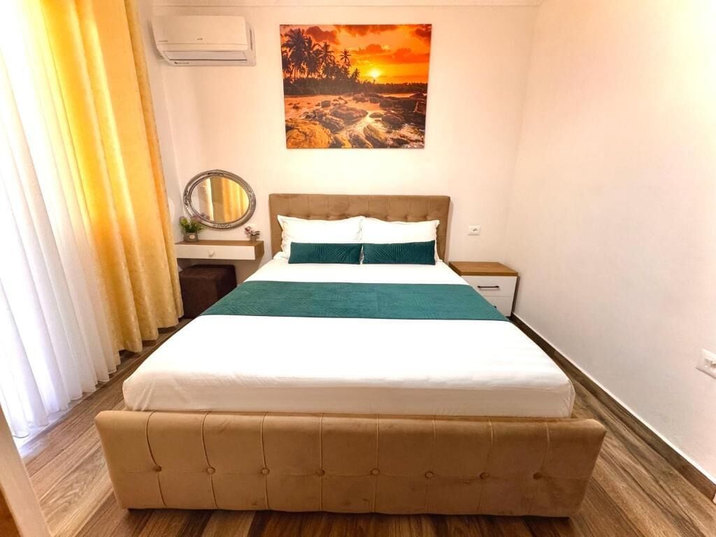 Une chambre d’hôtel comprenant un lit, une table de chevet, un miroir, un climatiseur fixé au mur et un tableau représentant un coucher de soleil.