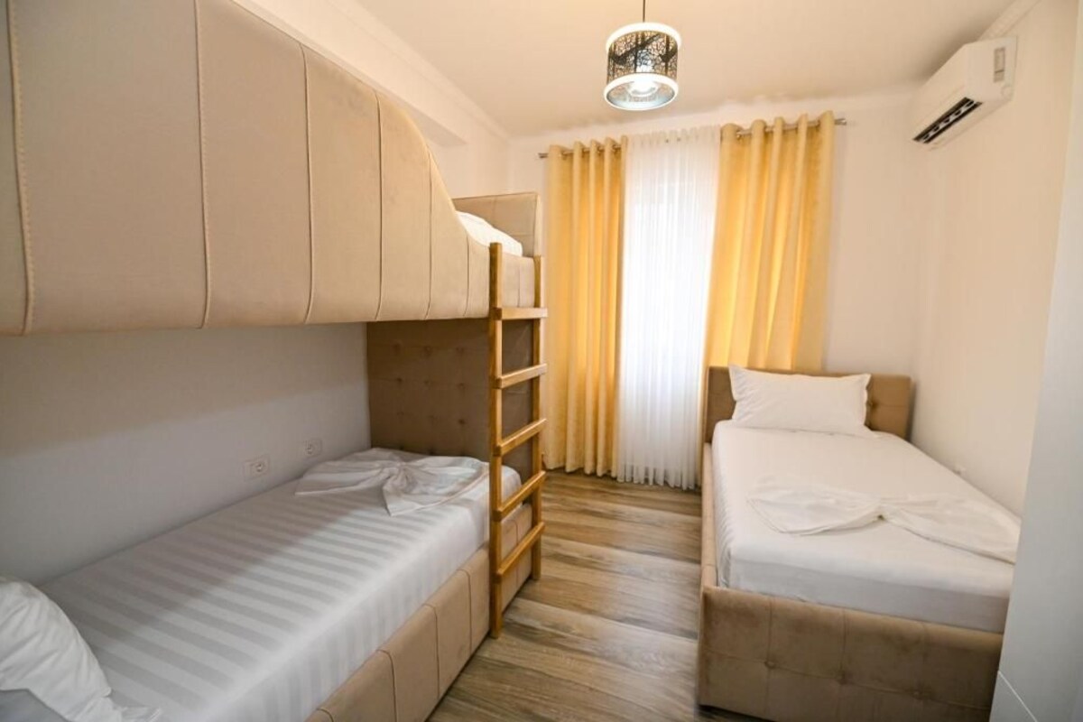 A compact hotel room with bunk beds, a wooden ladder, a small window with curtains, and a ceiling light fixture.