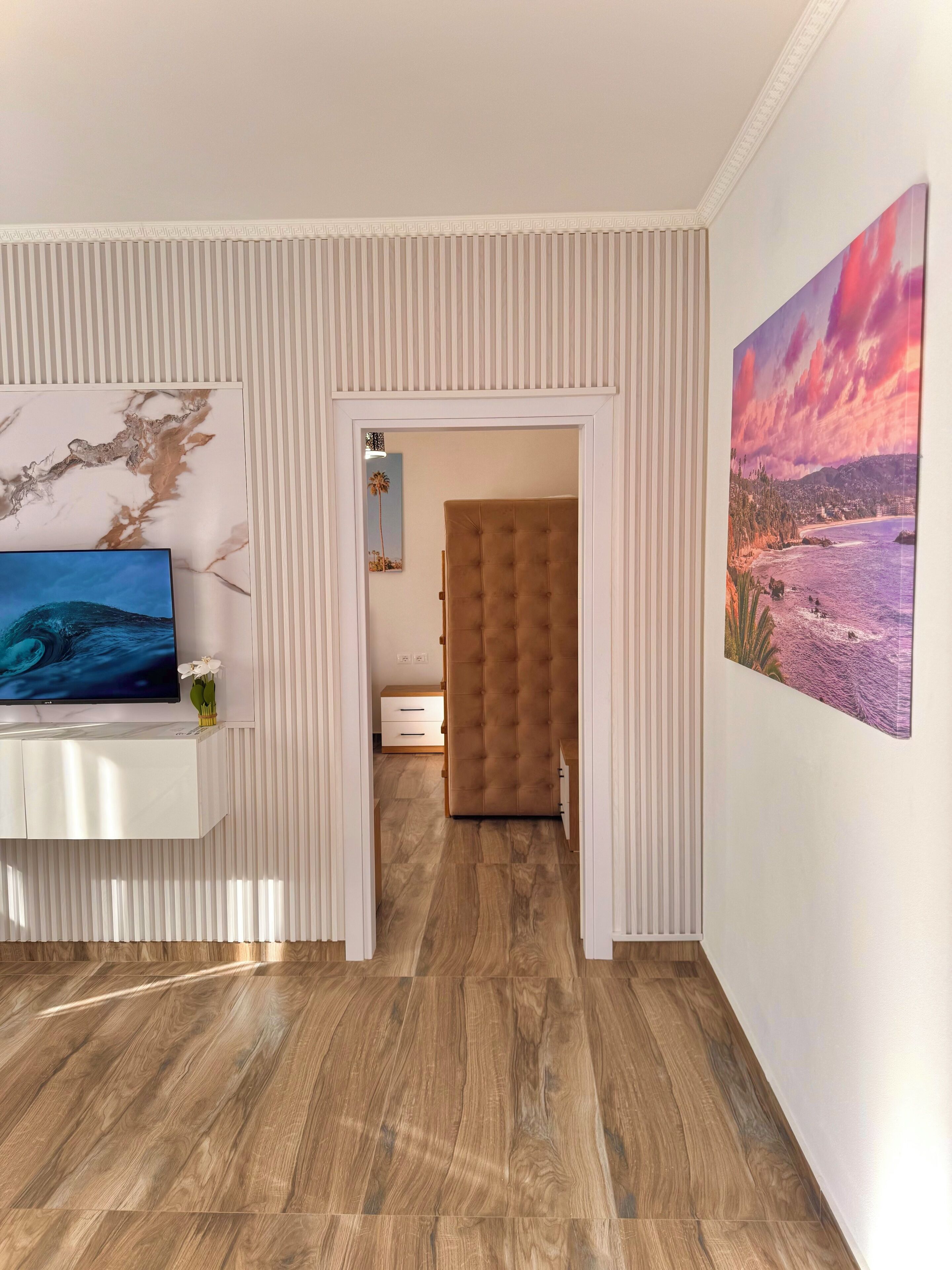 A modern interior with a wooden floor, a white console table with a TV, a tufted headboard, and a large landscape painting.