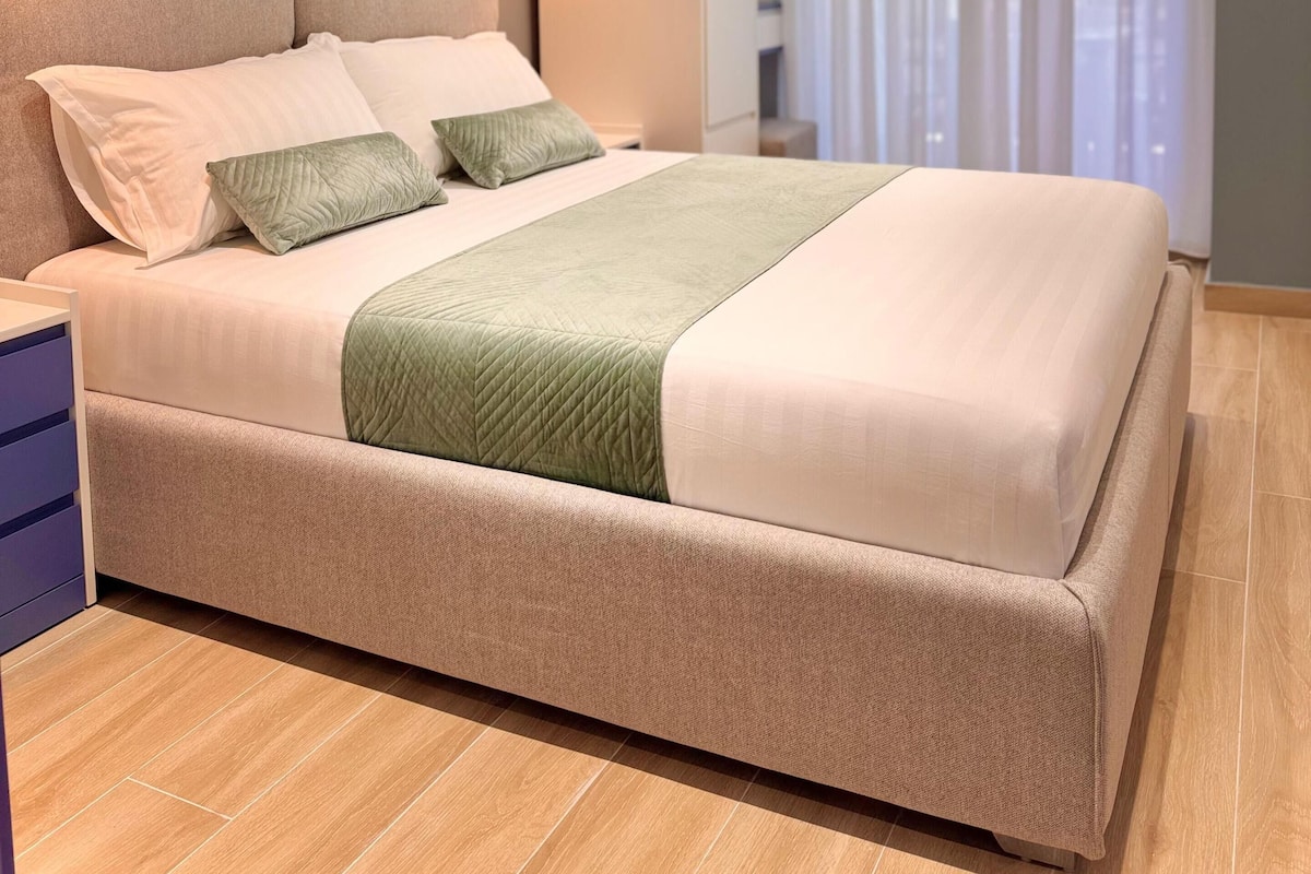 Un dormitorio moderno con una cama grande, una mesita de noche y vistas a la ciudad a través de cortinas translúcidas.