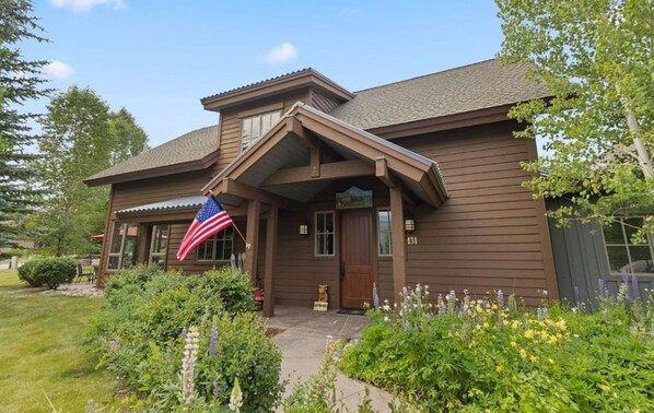 A two-story wooden house with a front porch, a flag, and a well-maintained garden.