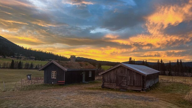 Una cabaña rústica con techo metálico y estructura de madera con techo inclinado, ambas ubicadas en un campo cubierto de hierba con el atardecer y montañas al fondo.