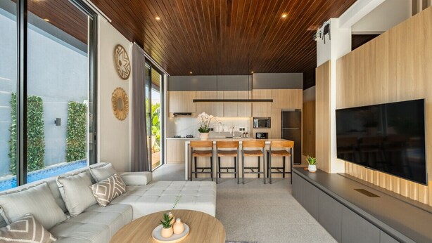 Uma cozinha moderna com ilha central, área de refeições com banquetas, uma TV grande na parede e um espaço de estar aconchegante com sofá e mesa de centro.