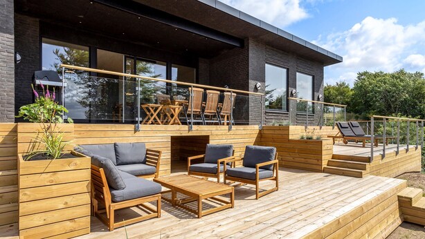 A modern outdoor deck with wooden furniture, a barbecue grill, and a glass railing overlooking a garden.