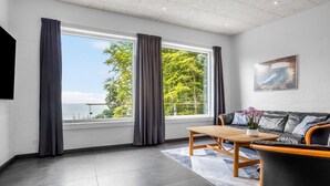 A modern living room with a large window, a leather sofa, a wooden coffee table, and a painting on the wall.