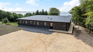 A modern, single-story building with a dark brick exterior and a flat roof, situated on a gravel lot with a wooden deck and a view of a body of water in the background.