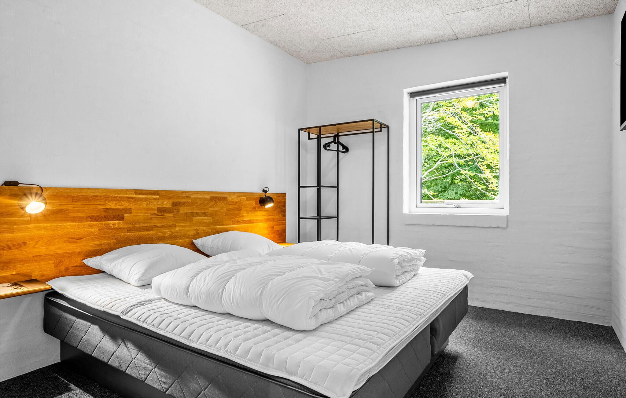 A bedroom with a wooden headboard, a bed with white bedding, a nightstand, a wall-mounted shelf, and a window with a view of greenery.