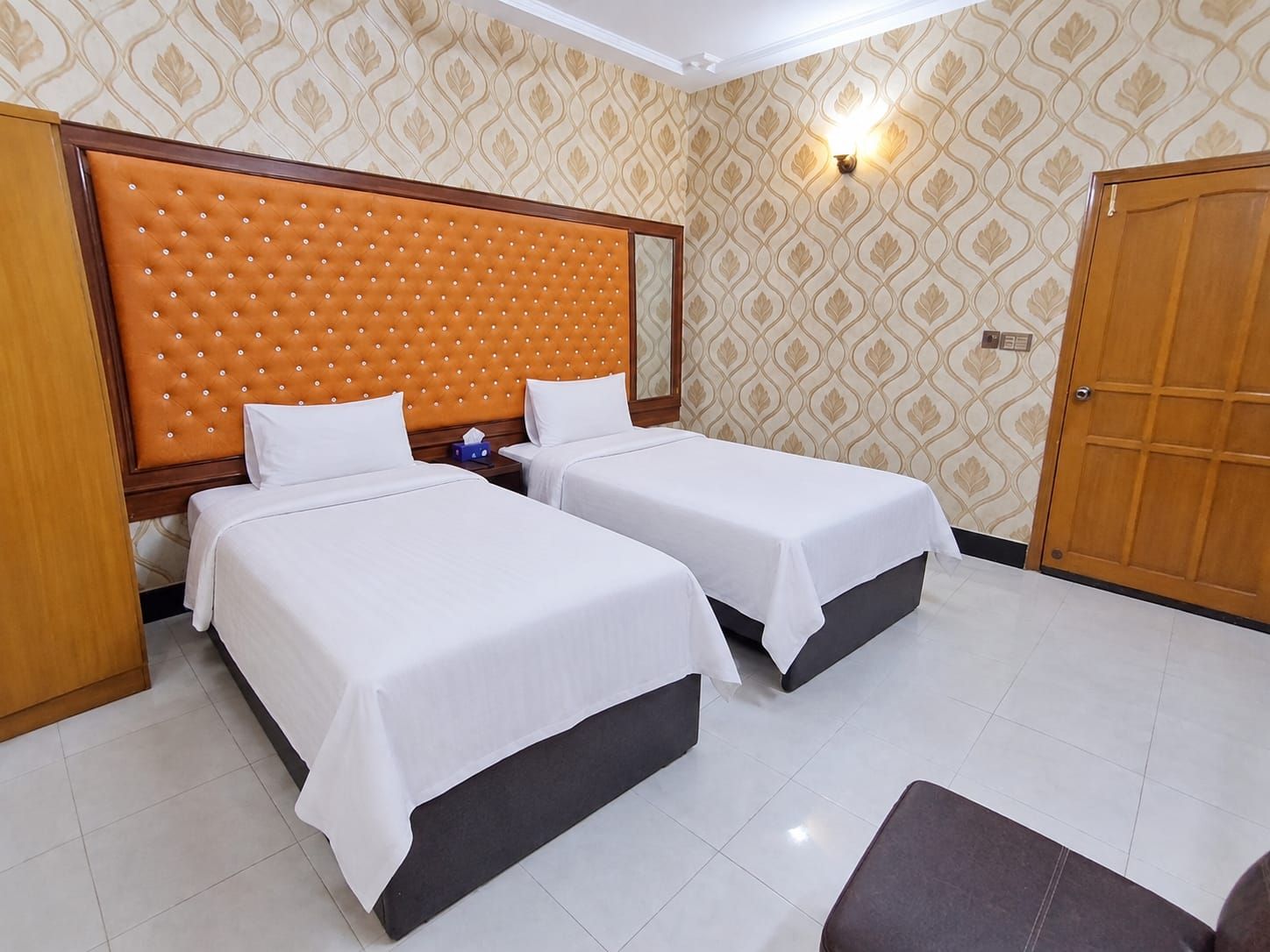 A hotel room with two beds, a wooden headboard, a nightstand, a wall-mounted light, and a door.