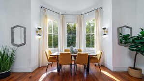 A dining area with a wooden table and chairs, large windows with curtains, and wall-mounted light fixtures. - Luxurious 4 Bedroom, 4.5 Bath Home (Charleston)