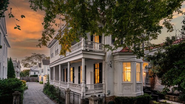 A white colonial-style house with a covered porch and a bay window, surrounded by greenery and a cobblestone path. - Luxurious 4 Bedroom, 4.5 Bath Home (Charleston)