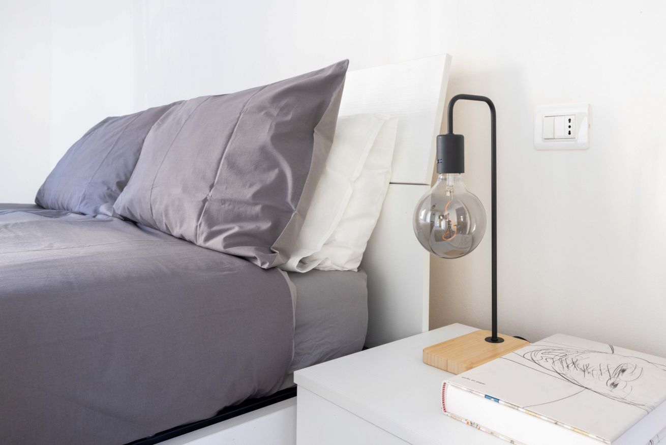 En pænt redt seng med gråt og hvidt sengetøj, et natbord med en lampe, og en stikkontakt på væggen.