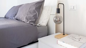 Una cama bien hecha con ropa de cama gris y blanca, una mesita de noche con lámpara y un enchufe en la pared.