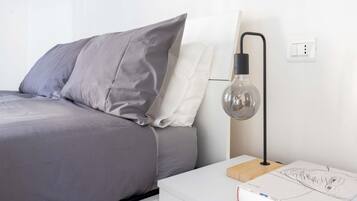 En pænt redt seng med gråt og hvidt sengetøj, et natbord med en lampe, og en stikkontakt på væggen.