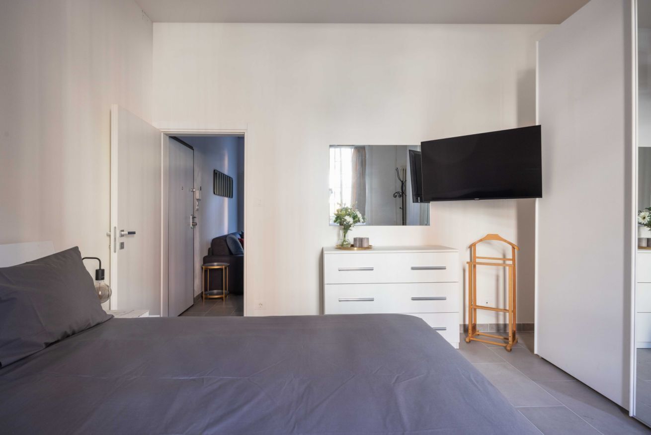 Une chambre à coucher moderne, dotée d’un grand lit, d’une commode, d’un téléviseur fixé au mur et d’une porte donnant sur une autre pièce.
