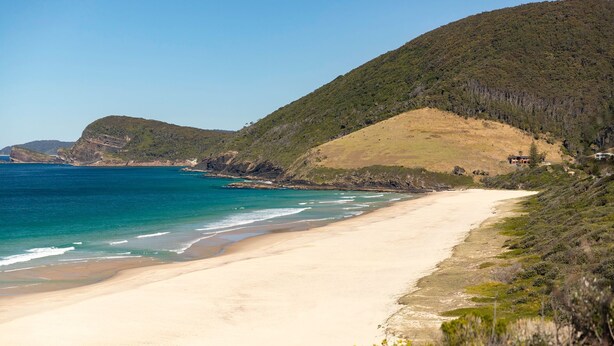 A coastal beach with a sandy shore, a clear blue sea, and a hilly backdrop.
