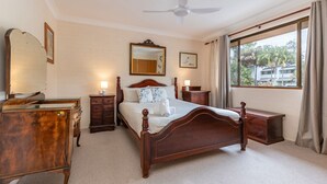 A bedroom with a wooden bed, a dresser, a nightstand, a mirror, and a window with a view of palm trees.