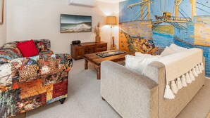 A cozy living room with a colorful sofa, a grey couch, a wooden coffee table, and a painting of a ship on a blue background.