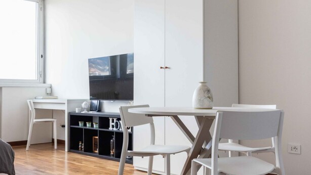 A modern living room with a white sofa, a dining table with white chairs, a flat-screen TV mounted on the wall, and a bookshelf with various items.