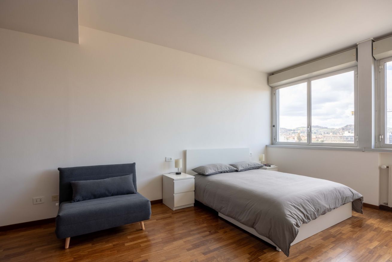 Un dormitorio con cama, mesita de noche, silla y ventana con vista a la ciudad.