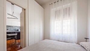 A bedroom with a large bed, a window with sheer curtains, and a view of a kitchen area.