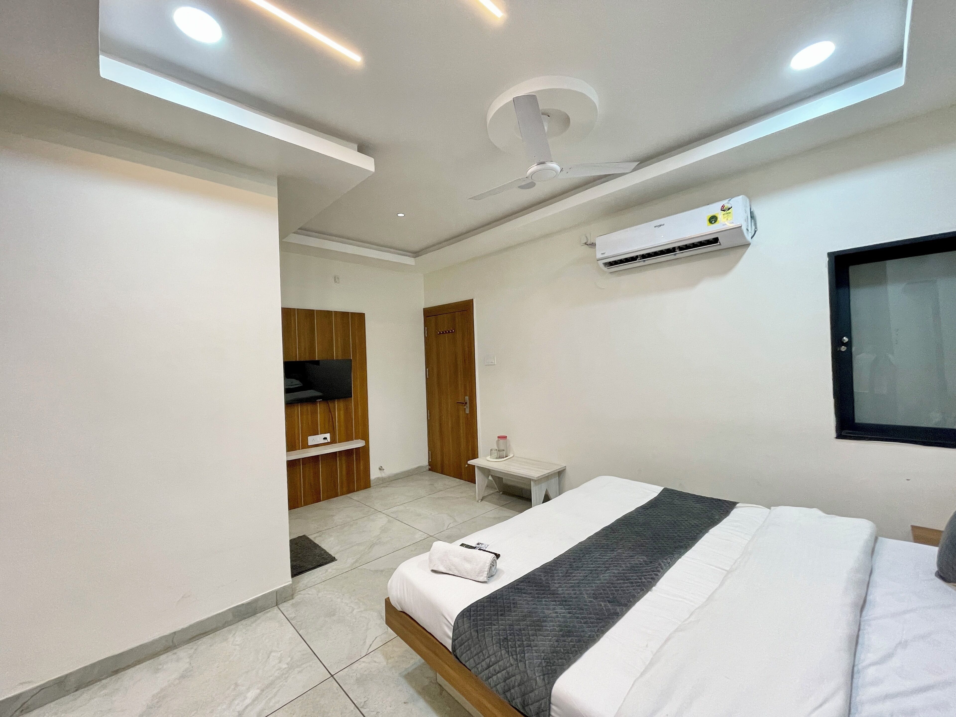 A modern hotel room with a bed, a small bedside table, a TV mounted on a wooden panel, an air conditioning unit, and a ceiling fan.