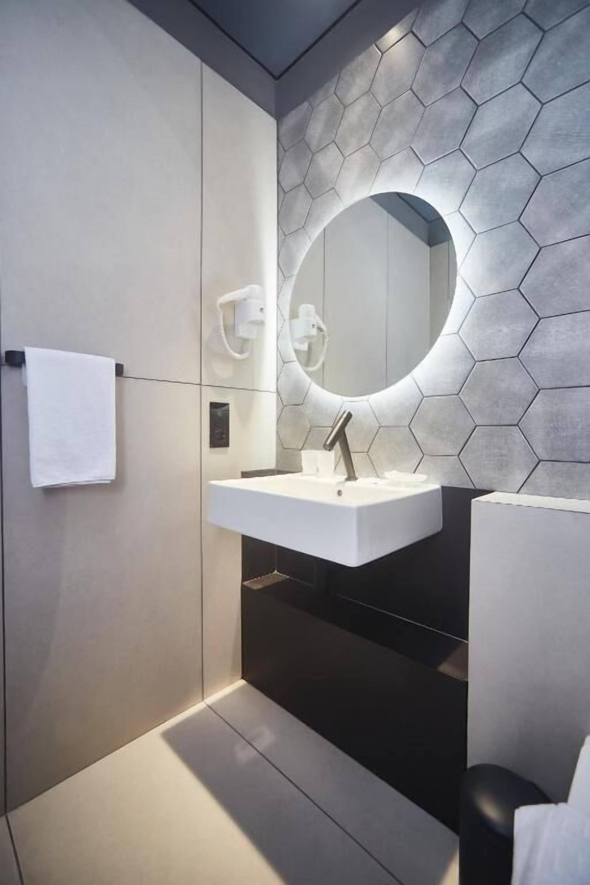 Un baño moderno con un lavabo blanco, un espejo redondo y un salpicadero de azulejos hexagonales.