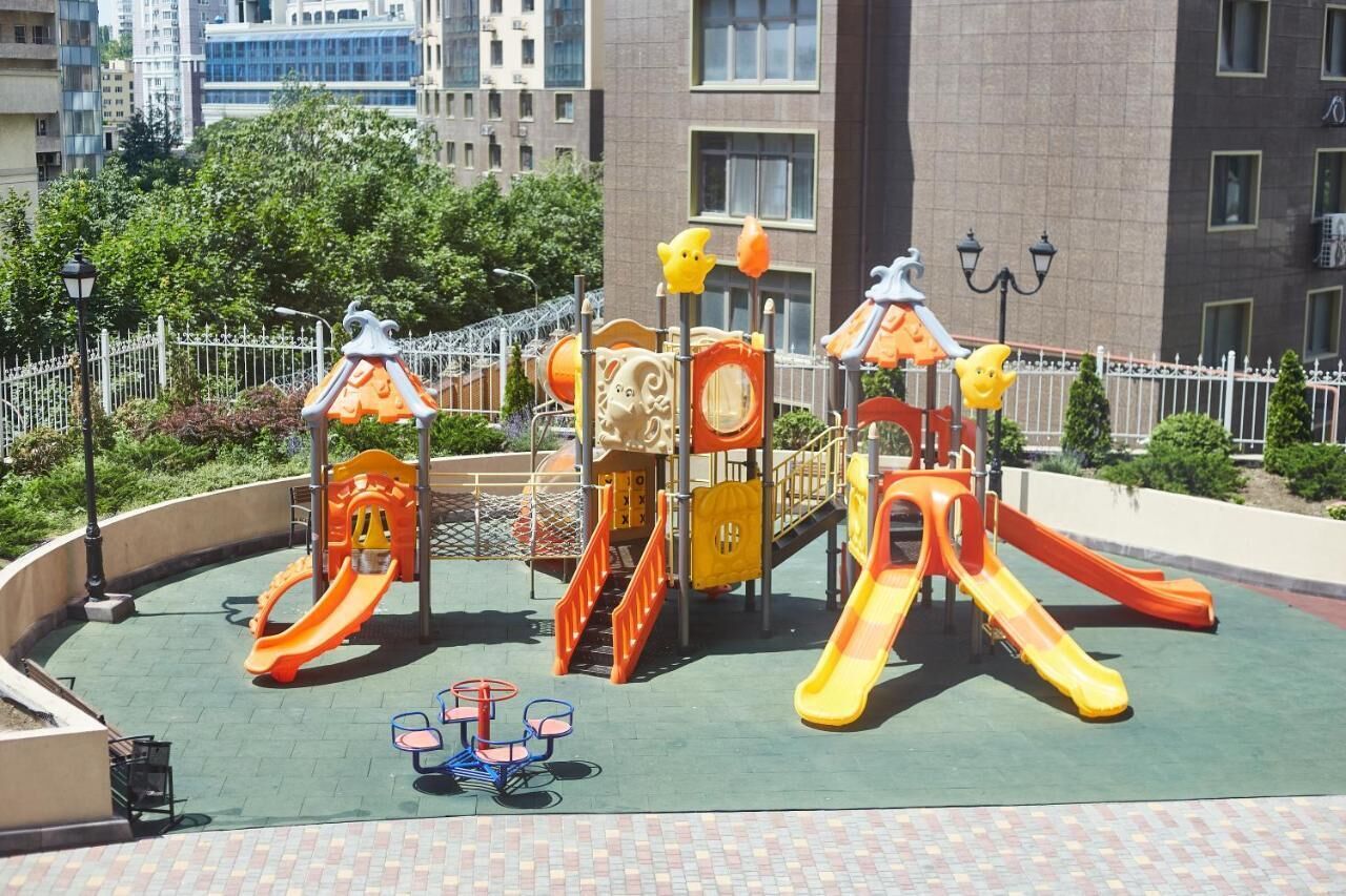A playground with slides and climbing structures, surrounded by a fence and buildings in the background.